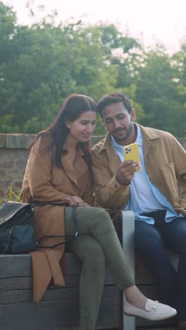 Vertical-Video-Of-Muslim-Couple-On-Date-Sitting-In-Outdoor-City-Park-Looking-At-Mobile-Phone-Together